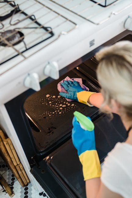 Close-up of a person wearing a yellow and blue rubber glove, using a blue cloth to clean the black glass surface of a kitchen oven. The oven's glass door is slightly open, revealing the interior racks