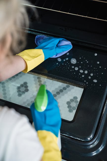 Close-up of a person cleaning the oven's glass door with a blue and yellow rubber glove, using a blue cloth to wipe away soap suds and grease. The oven's interior, visible through the glass, shows a d
