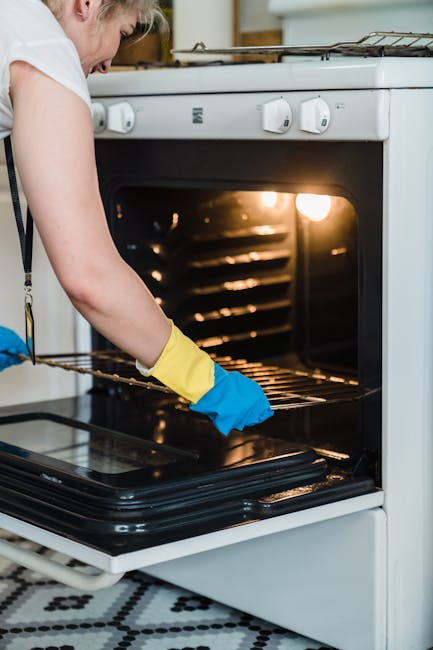 Close-up of a person cleaning the oven's glass door with a blue and yellow rubber glove, using a blue cloth to wipe away soap suds and grease. The oven's interior, visible through the glass, shows a d